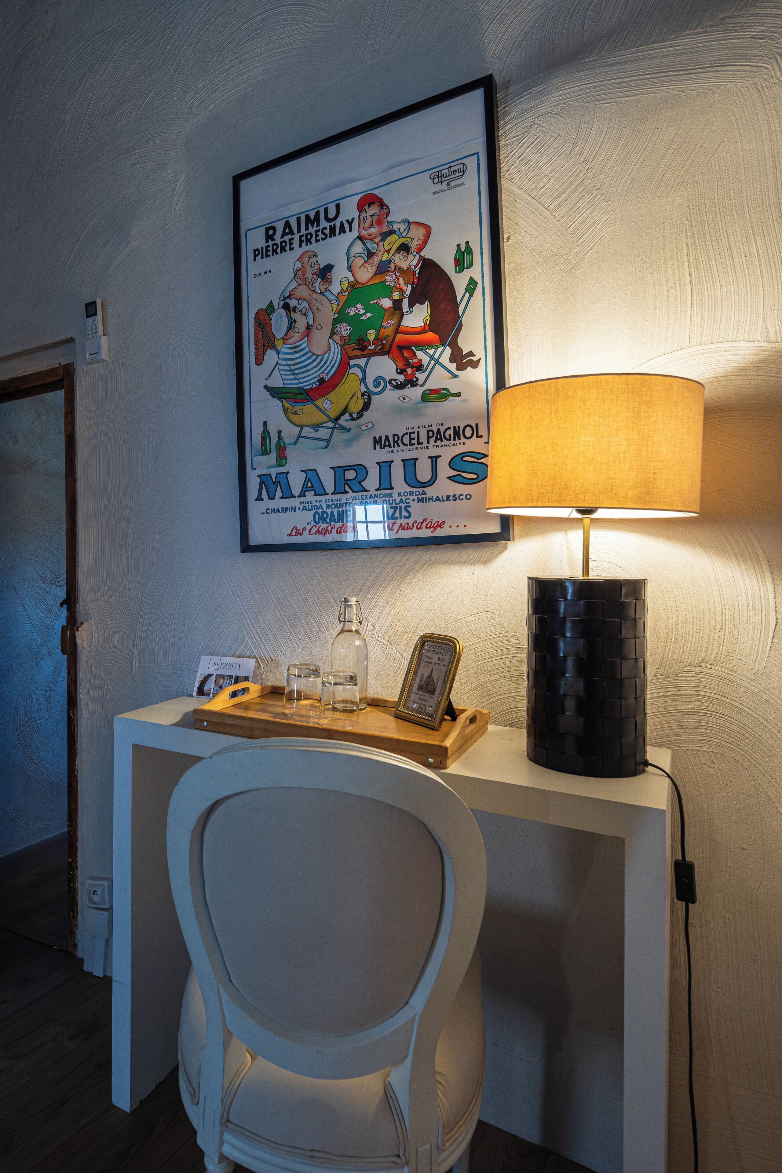 A room with a framed movie poster on the wall, a desk with a lamp, a bottle, and glasses.
