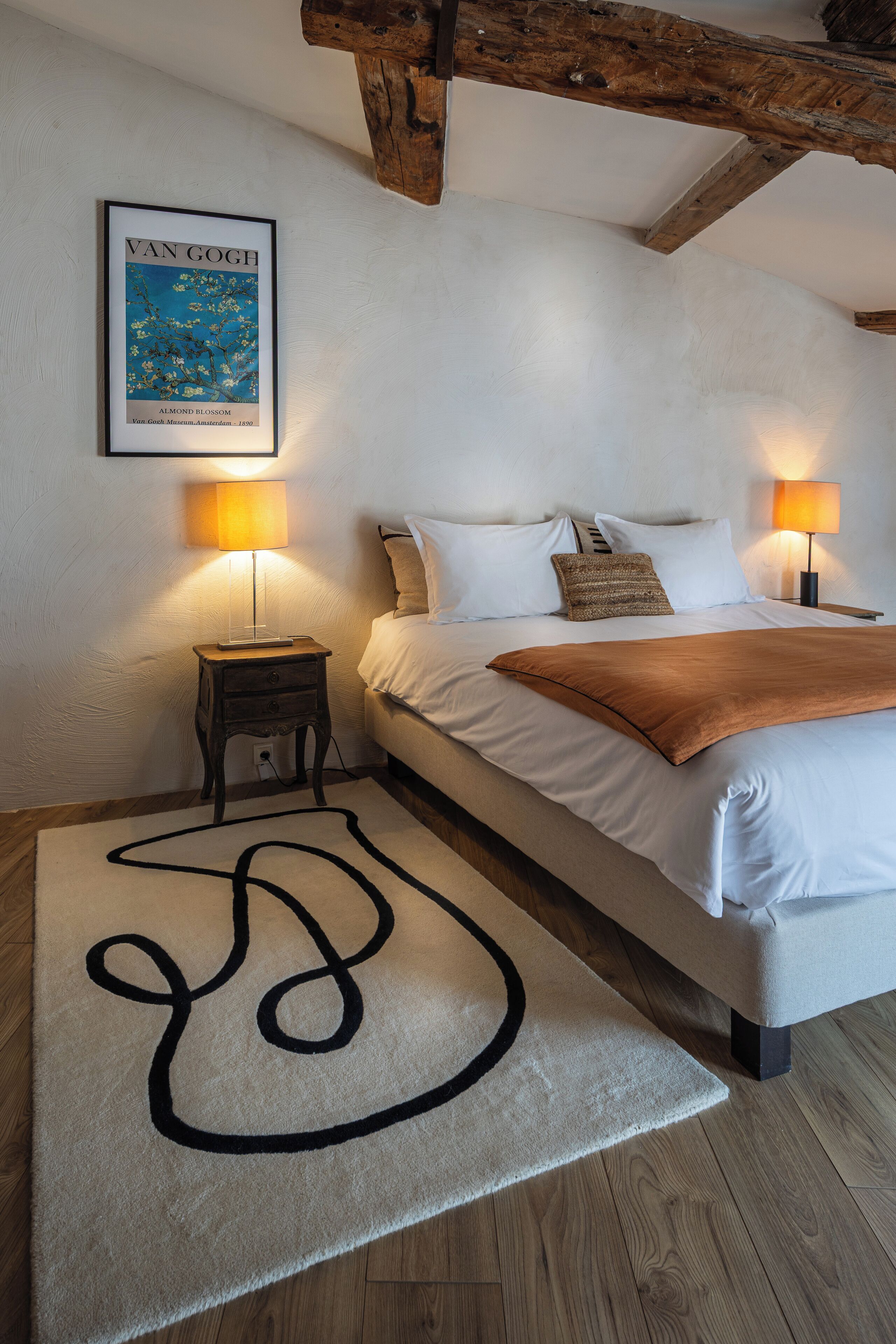 Un dormitorio con una cama, dos lámparas de noche, una obra de arte enmarcada en la pared y una alfombra de diseño sencillo.