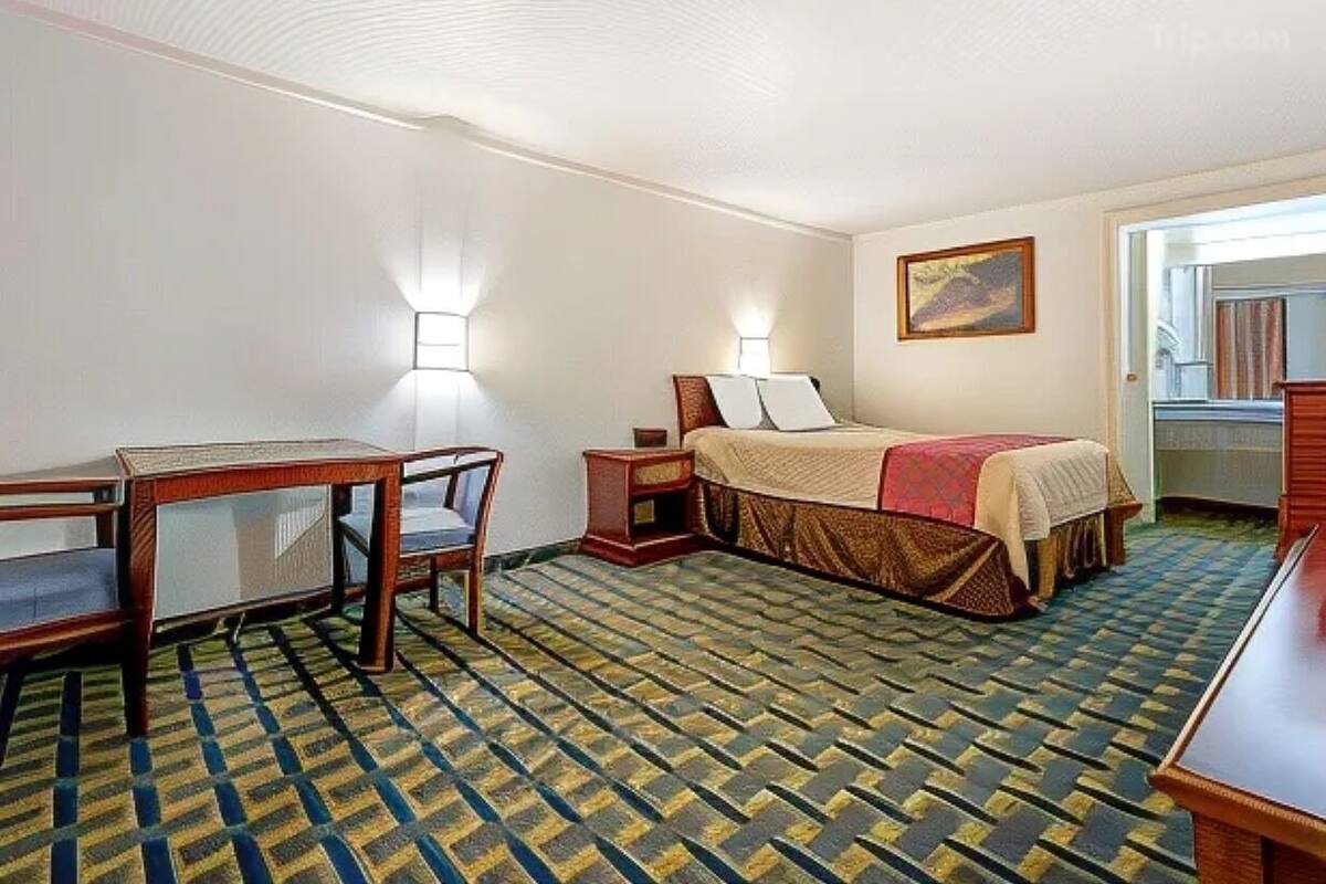A hotel room with a bed, a desk, and a chair. There are two wall-mounted lamps and a painting on the wall.