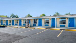 A motel with a row of rooms, each with a blue door and a window. There is a parking lot in front of the motel. - Hilliard Motor Lodge (Hilliard)