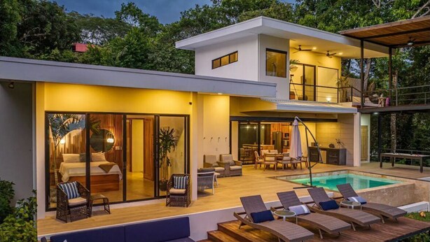 Ein modernes Haus mit zwei Etagen, einem großen Außenpool, Liegestühlen und einer Feuerstelle.