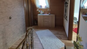 Un couloir avec un escalier en bois, un tapis blanc, une petite fenêtre avec des rideaux et un meuble contenant divers objets.