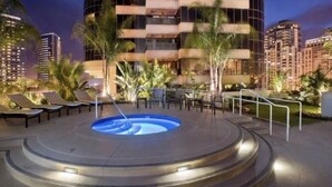 A pool area with lounge chairs and a circular hot tub, surrounded by palm trees and modern buildings in the background.