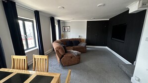 Een moderne woonkamer met een bruine bank, houten stoelen, een eettafel en een groot televisiescherm.