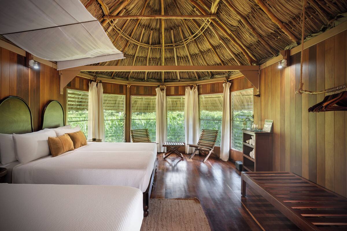 A room with a bed, a wooden bench, and a table. There are windows with curtains, and a thatched roof ceiling.