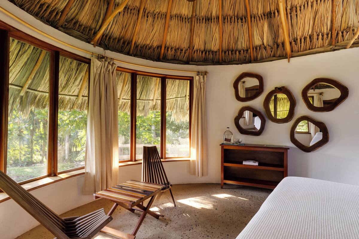 A room with a thatched roof, large windows, a wooden chair, a bed, and several mirrors on the wall.