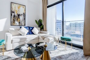 A modern living room with a white sofa, a glass coffee table, and a view of the city through large windows. - Luxe Gardenia 2BR Haven in Dubai (Dubai)