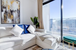 A modern living room with a white sofa, a large abstract painting, and a view of a cityscape through a large window. - Luxe Gardenia 2BR Haven in Dubai (Dubai)