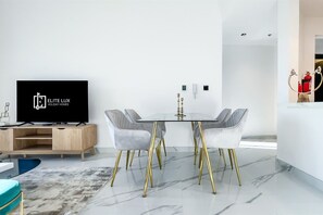 A modern living room with a flat-screen TV, a wooden entertainment unit, a glass dining table with gold legs, grey upholstered chairs, and a marble floor. - Luxe Gardenia 2BR Haven in Dubai (Dubai)