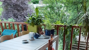 Terraza de madera con una mesa y sillas blancas, rodeada de plantas en macetas y vistas al verdor.