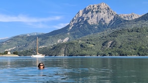 Una persona nada en un lago con un velero y montañas al fondo.