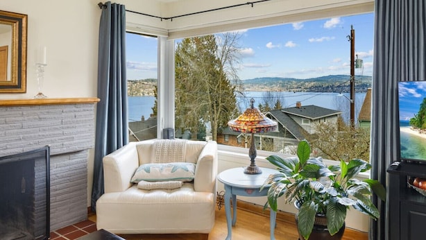 A living room with a white sofa, a small table with a lamp, and a large window offering a view of a lake and buildings. - Lake Washington View Home with Hot Tub BBQ Garden (Seattle)