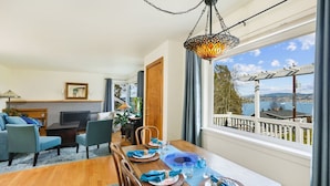 A dining area with a wooden table set for four, a TV in the background, and a view of a lake through large windows. - Lake Washington View Home with Hot Tub BBQ Garden (Seattle)