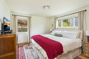Una camera da letto con un letto, una televisione su una cassettiera in legno, una finestra con tende e vista su un edificio.