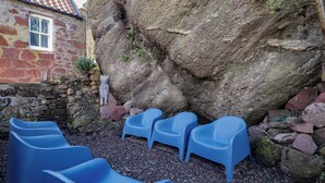Un edificio de piedra con techo de tejas, una estatua y sillas de plástico azules dispuestas al aire libre.