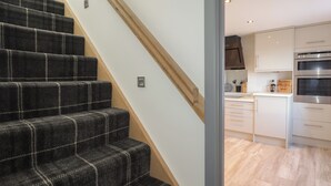 Una escalera con alfombra a cuadros oscura lleva a una zona de cocina con armarios blancos y electrodomésticos de acero inoxidable.