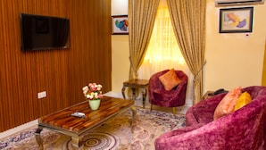 Una sala de estar con pared de paneles de madera, un televisor de pantalla plana montado en ella, una mesa de centro con un jarrón de flores, una alfombra estampada y dos sillones morados.