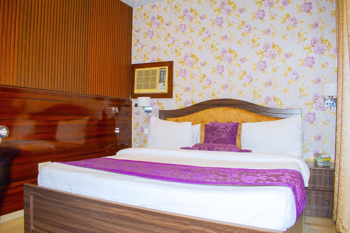 A neatly made bed with a wooden headboard, a floral wallpaper pattern, and a small bedside table with a lamp.