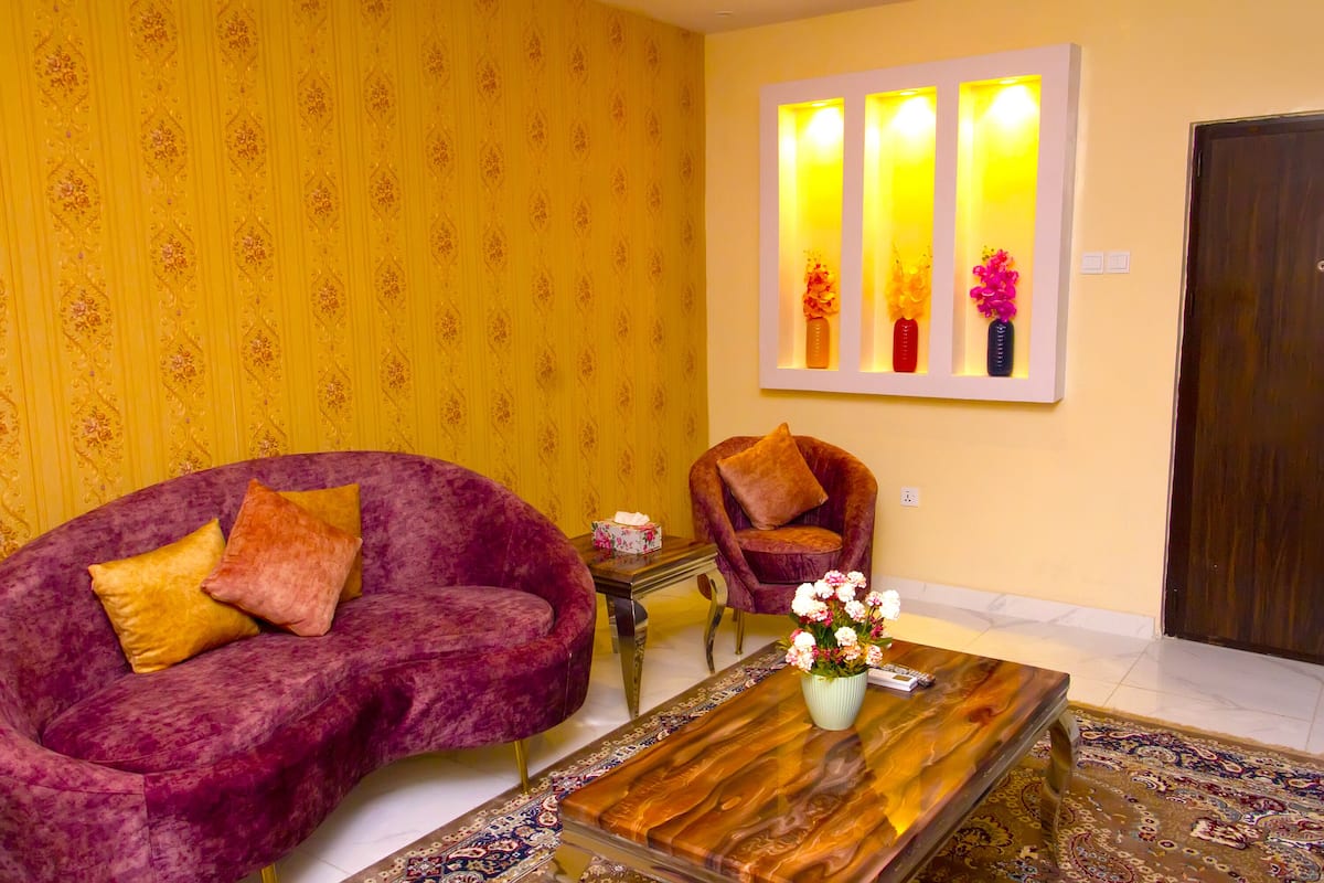 A living room with a purple sofa, a wooden coffee table with a vase of flowers, and a wall-mounted light fixture.