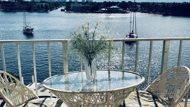Un balcón con una mesa de vidrio y sillas de mimbre, con vista a una masa de agua con barcos y el perfil de una ciudad en la distancia.
