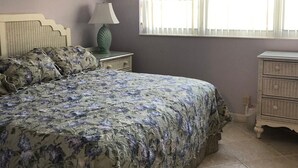 Un dormitorio con una cama grande, una mesita de noche con lámpara, un aparador y una ventana con persianas.