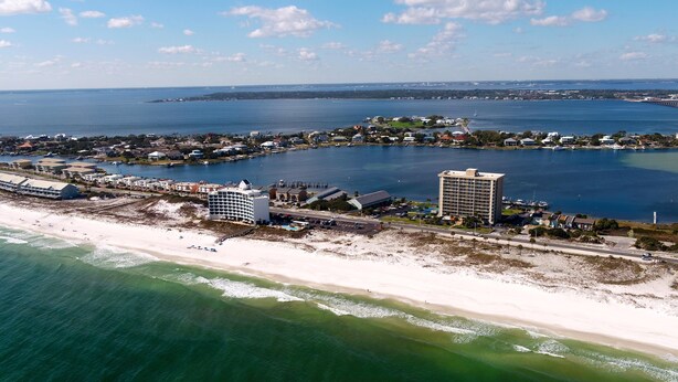 An aerial view of a coastal area with a beach, a marina, and several buildings including a hotel with a large pool and a tall hotel building. - Chateau by the Bay, Close to Restaurants and Bars, Walk to the Gulf or Bay- W209 (Pensacola Beach)