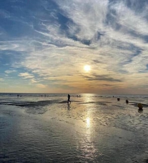 Une plage où l’on voit des gens se promener, le soleil se coucher et un ciel dégagé.