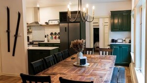 Un coin repas avec une table en bois, des chaises et un lustre. On aperçoit au fond une cuisine avec des meubles verts et un réfrigérateur.