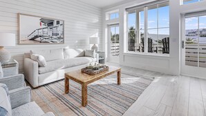 A modern living room with a white sofa, a wooden coffee table, and large windows offering a view of the outdoors. - She Sells Seashells TC 8203 (Port Aransas)
