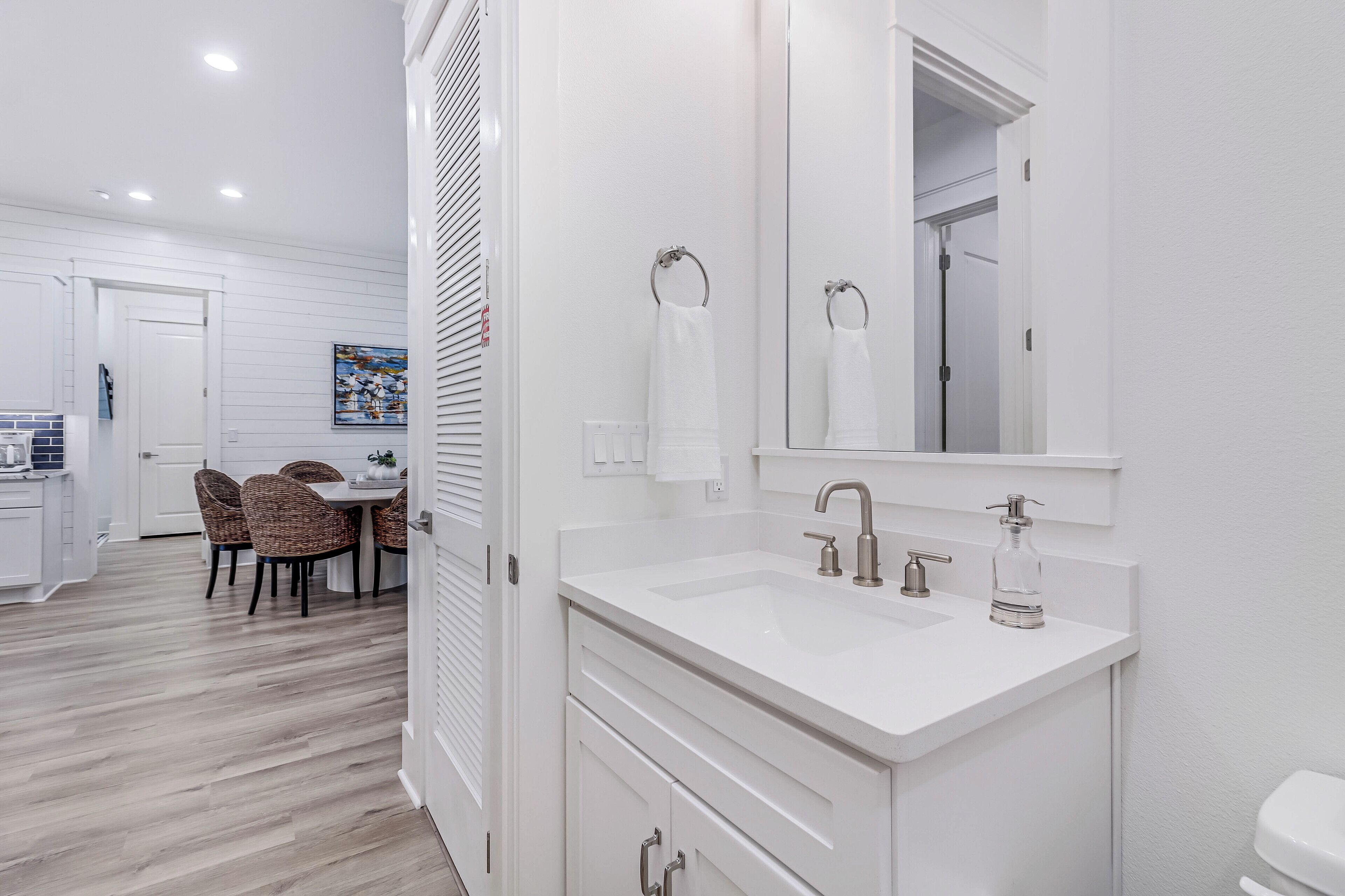 A modern bathroom with a white sink, mirror, and toilet. There is a dining area with wicker chairs and a table visible in the background.