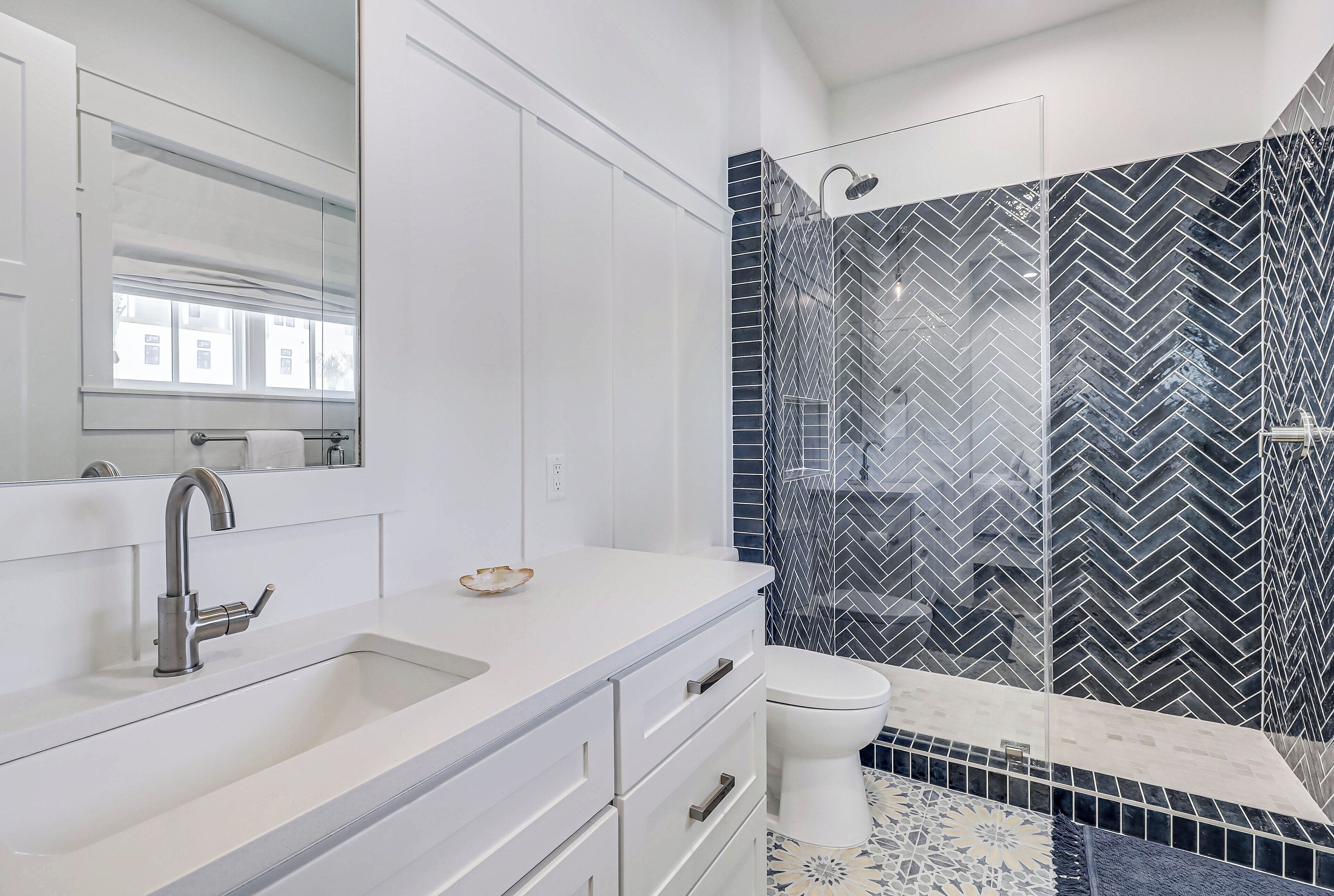 A modern bathroom with a white vanity, a sink with a faucet, a toilet, and a shower area with a black and white herringbone tile backsplash.