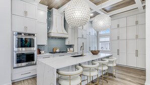 Una cocina moderna con armarios blancos, una isla central con taburetes de bar, electrodomésticos de acero inoxidable e iluminación colgante hexagonal.