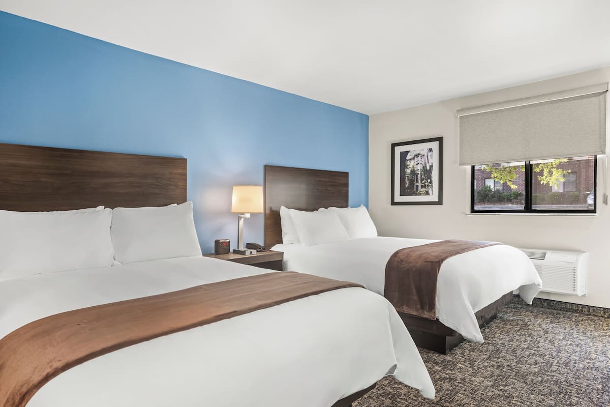 A hotel room with two beds, a nightstand with a lamp, a window with a view, and a framed picture on the wall.