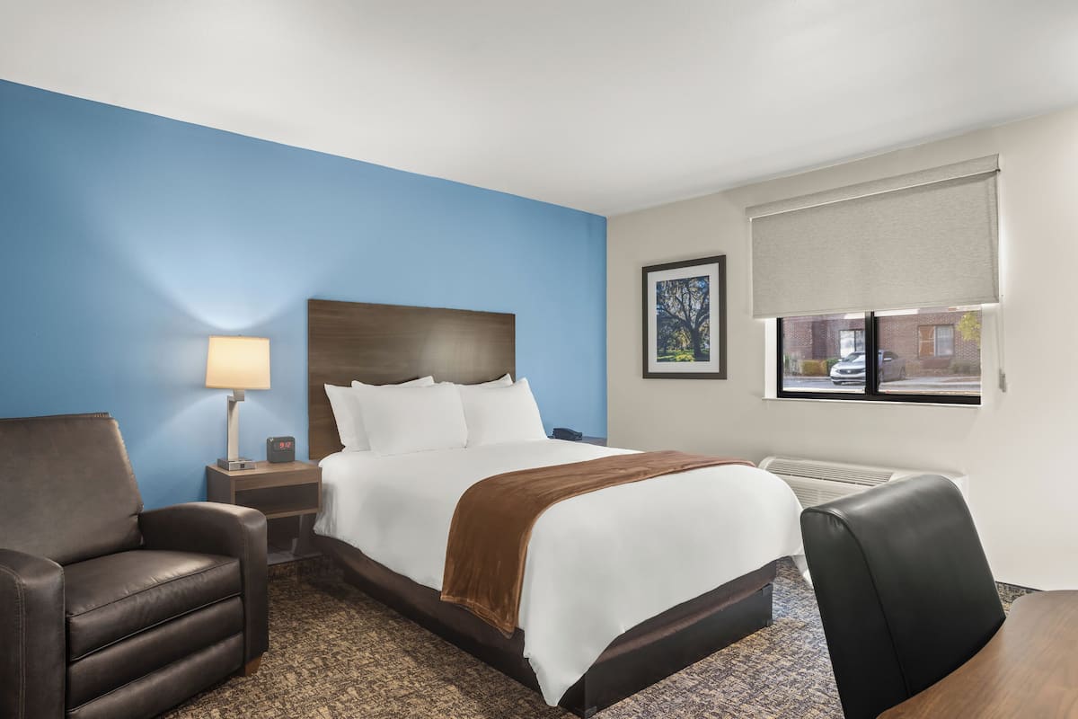 A hotel room with a bed, a desk, a chair, a lamp, and a window with a view of a building.