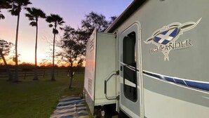 A highlander camper with a door open, a blue-striped pathway leading to it, and palm trees in the background. - Sunniland Ranch Airport 08FD Fly-In Destination

 (Okeechobee)