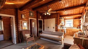 Interior de una cabaña de troncos con sofá, sillón y una mesa de centro en la sala de estar, armarios de madera en la cocina y un ventilador de techo.