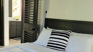 Ein ordentlich bezogenes Bett mit weißer Bettwäsche und gestreiften Kissen, ein hölzernes Kopfteil sowie Blick auf ein Badezimmer mit dunkler Tür und Fenster.