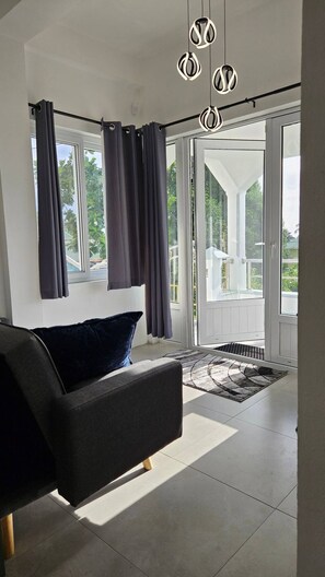 A modern living room with a sofa, a large window with curtains, and a view of greenery outside. - Village House 2 (Vieux Fort)