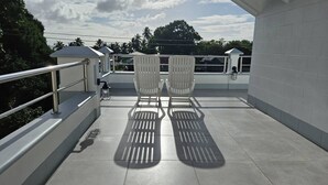 Eine Dachterrasse mit zwei weißen Stühlen und einem Tisch, einem Metallgeländer und Blick auf Palmen und Gebäude.