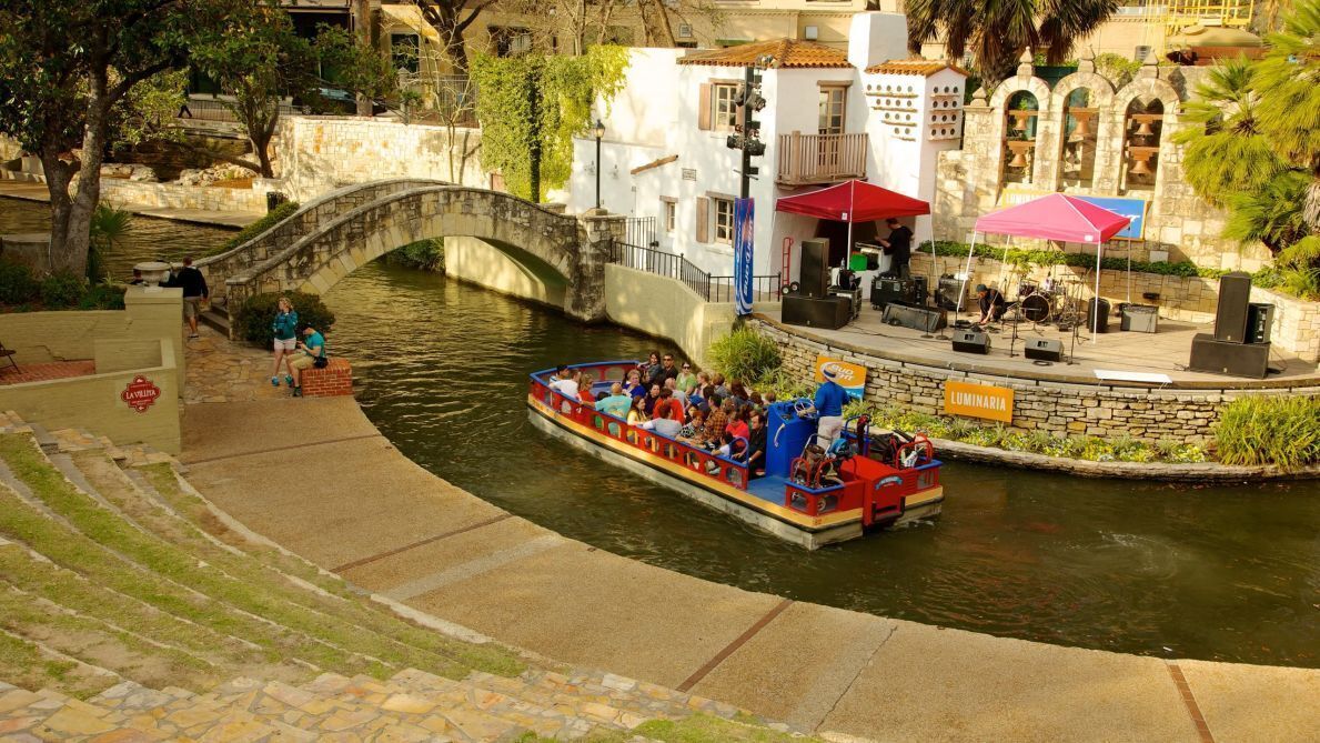 Un recorrido en barco por un canal con un puente de piedra, un restaurante con terraza y un letrero que dice 'LUMINARIA'.