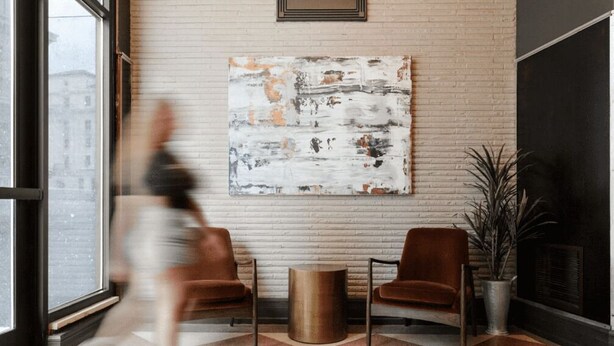 Un vestíbulo moderno con un cuadro abstracto grande en la pared, dos sillones marrones, una mesa redonda pequeña y una planta en maceta.