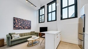 A modern living room with a sofa, a coffee table, a TV, and a kitchen area with a fridge and cabinets.