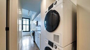 A laundry room with stacked washing machines and dryers, a kitchen area with white cabinets, and a large window providing natural light.