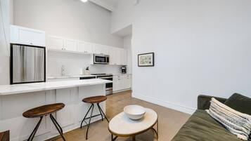 A modern kitchen with white cabinets, a stainless steel refrigerator, a microwave, and a coffee machine. There are two stools and a small round table with a bowl. A green couch is visible in the foreground.