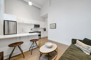 Una cocina moderna con armarios blancos, un refrigerador de acero inoxidable, un microondas y una máquina de café. Hay dos taburetes y una pequeña mesa redonda con un tazón. Se ve un sofá verde en primer plano.