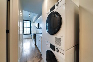 A laundry room with stacked washing machines and dryers, a kitchen area with white cabinets, and a large window providing natural light. - Historic Light, Modern Living at La Luz (San Antonio)