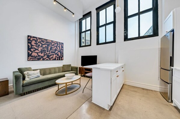 Un salon moderne comprenant un canapé, une table basse, un téléviseur et un coin cuisine avec un réfrigérateur et des placards.