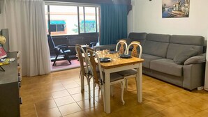 Un salon avec un canapé gris, une table à manger en bois avec des chaises, et une vue sur l’extérieur par une fenêtre.