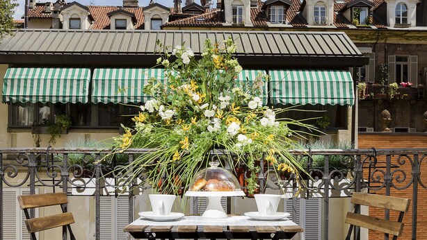 Un balcon avec une table dressée pour deux personnes, entouré de chaises en fer forgé, d’une plante en pot et d’un arrangement floral.