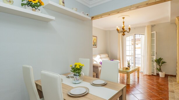 A dining area with a wooden table, white chairs, and a vase of yellow flowers. There is a chandelier above the table and a view of a living room in the background.